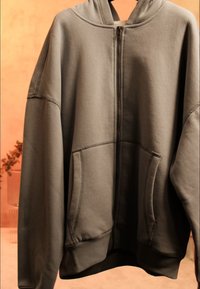 Oversized gray zip-up hoodie with front pockets and visible seam details, hanging against a blurred warm-toned background.