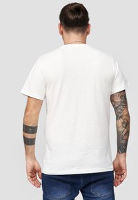 Vit bomullströja med korta ärmar, rundad halsringning och en enkel baksida. Har tatueringar på underarmarna och mörkblå jeans.