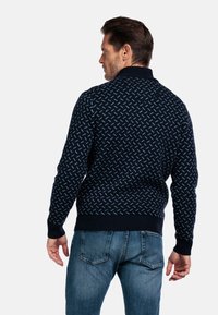 Marineblauwe gebreide trui met een geometrisch patroon in lichtblauw, geribbelde boorden en zoom, en een klassieke kraag. Gedragen over blauwe jeans.