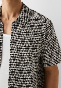 Persona vistiendo una camisa de botones de manga corta con un patrón abstracto en negro y beige sobre una camiseta sin mangas blanca con canalé.