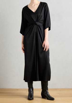 Femme portant une robe midi noire avec un nœud devant, manches trois-quarts, et des bottes noires à talons mi-mollet, debout sur un sol en bois.