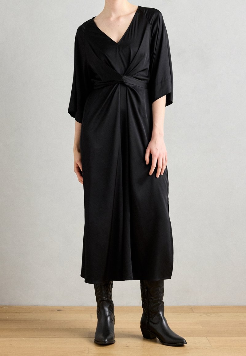 Femme portant une robe midi noire avec un nœud devant, manches trois-quarts, et des bottes noires à talons mi-mollet, debout sur un sol en bois.