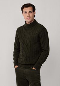 Man som bär en mörkgrön kabelstickad polotröja och matchande byxor med händerna i fickorna, stående mot en neutral bakgrund.