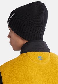 Svart ribbestrikket lue med "Timberland"-logo. Gul fleecejakke med teksturert overflate og blå tre-logo på baksiden av kragen.