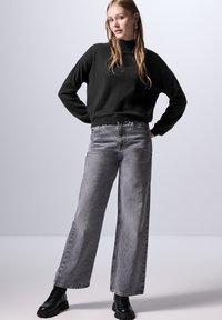 Maglione nero a maglia con colletto alto, abbinato a jeans grigi a gamba larga. Tessuto dalla texture liscia e stivaletti neri massicci.