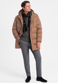 Giacca a vento color marrone chiaro con cappuccio, maglione grigio, pantaloni grigi e sciarpa nera. Stivali neri alla caviglia completano l'outfit.