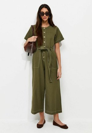 Femme portant une combinaison verte olive boutonnée avec ceinture, portant un sac à bandoulière marron, des lunettes de soleil foncées et des ballerines marron, debout contre un mur blanc.