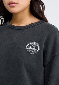 Felpa nera con scollatura consumata, caratterizzata da un logo a cuore ricamato bianco che recita "Buffy l'ammazzavampiri."