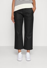 Pantalones de pierna ancha negros, confeccionados con una tela resistente, que presentan bolsillos frontales y un corte recto, combinados con zapatillas blancas sobre un fondo claro.