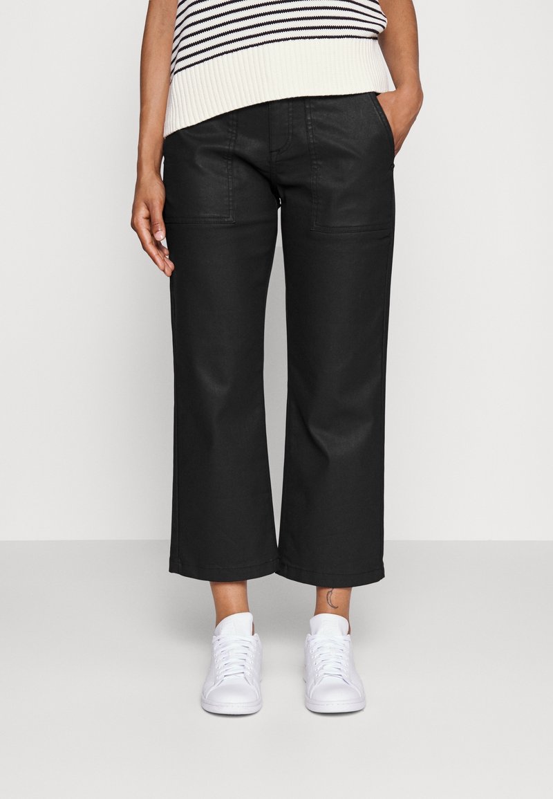 Pantalones de pierna ancha negros, confeccionados con una tela resistente, que presentan bolsillos frontales y un corte recto, combinados con zapatillas blancas sobre un fondo claro.