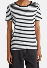 Vit kortärmad t-shirt med smala svarta ränder, svart rund halsringning och en subtil logotypdetalj nära fållen.