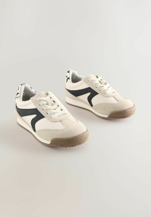 Par bež i bijelih tenisica s crnim bočnim detaljima i smeđim teksturiranim potplatima na svijetloj neutralnoj pozadini.