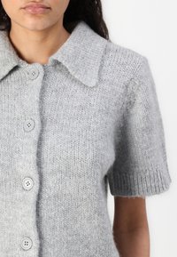 Šedý pletený cardigan s krátkým rukávem vyrobený z měkké vlny, s límečkem a pěti knoflíky. Texturovaná látka s ribovanými manžetami.