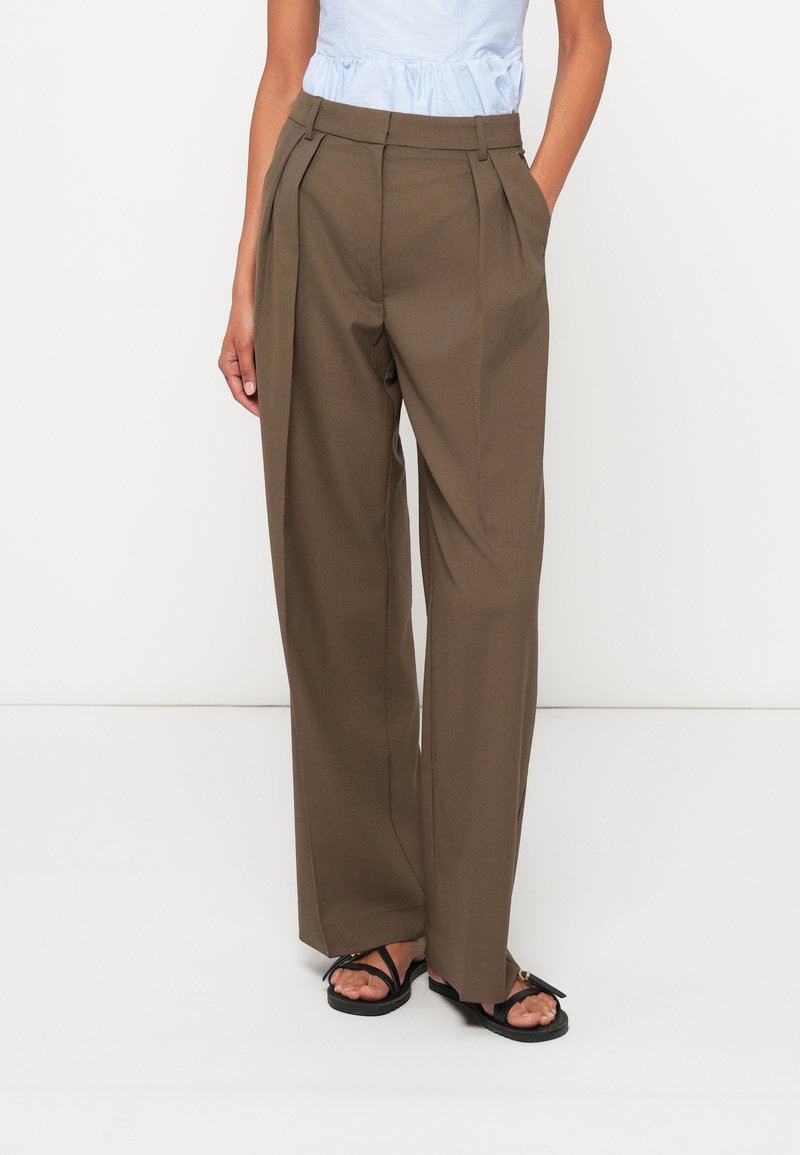 Hnedé plisované nohavice s vysokým pásom, širokými nohavicami a hladkou textúrou. Skombinované s čiernymi sandálmi a svetlomodrým topom.