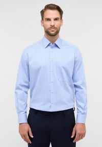 Camicia azzurro chiaro a maniche lunghe con righe verticali bianche, con bottoni davanti, colletto a punta e una tasca sul petto, abbinata a pantaloni scuri.