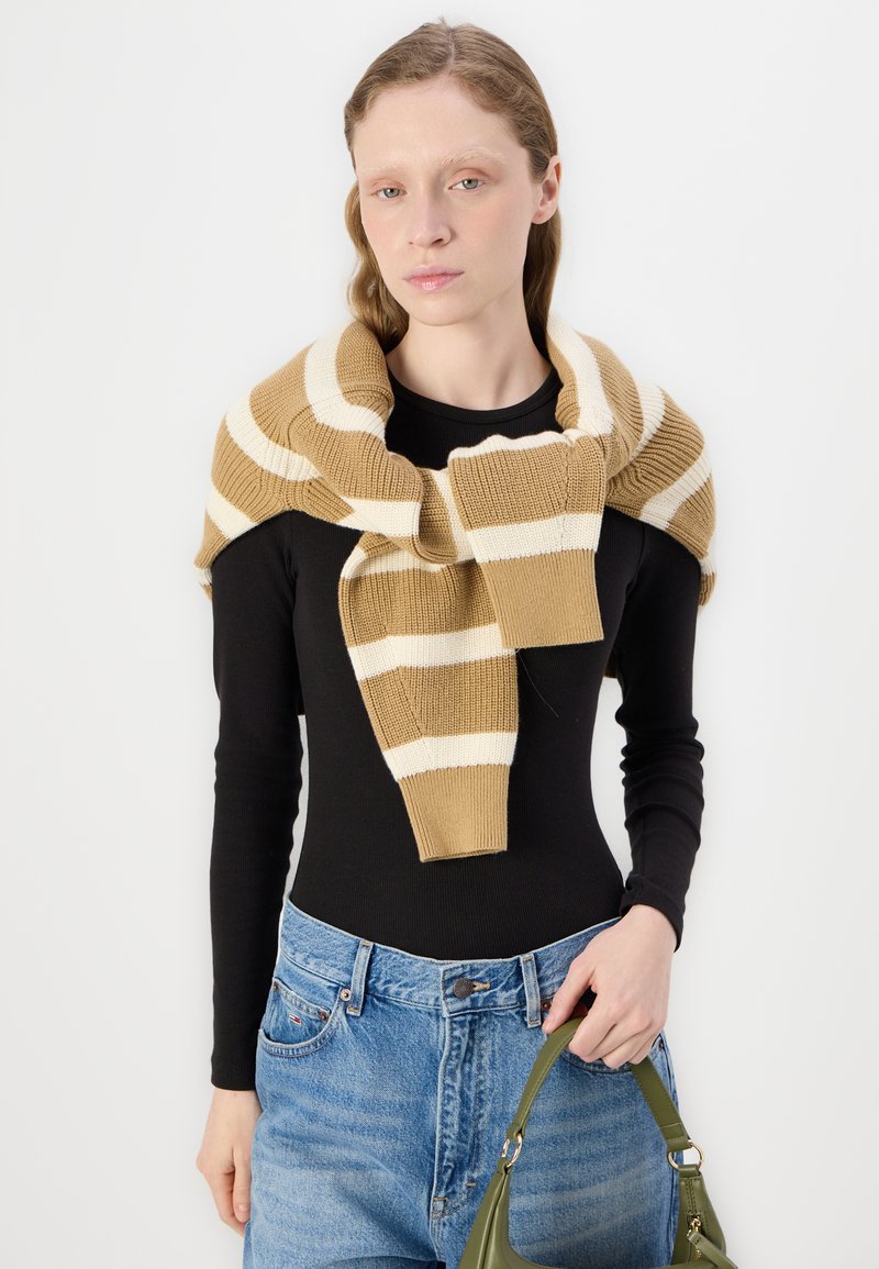Pull en tricot avec des rayures beiges et blanches, porté par-dessus un haut à manches longues noir. Assorti avec un jean taille haute bleu clair et un sac vert.