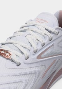 Λευκό αθλητικό παπούτσι Reebok με υφασμάτινη υφή, λευκά κορδόνια, ροζ χρυσή ετικέτα της μάρκας στα κορδόνια και ροζ χρυσές λεπτομέρειες κατά μήκος του πλαισίου και της γλώσσας.