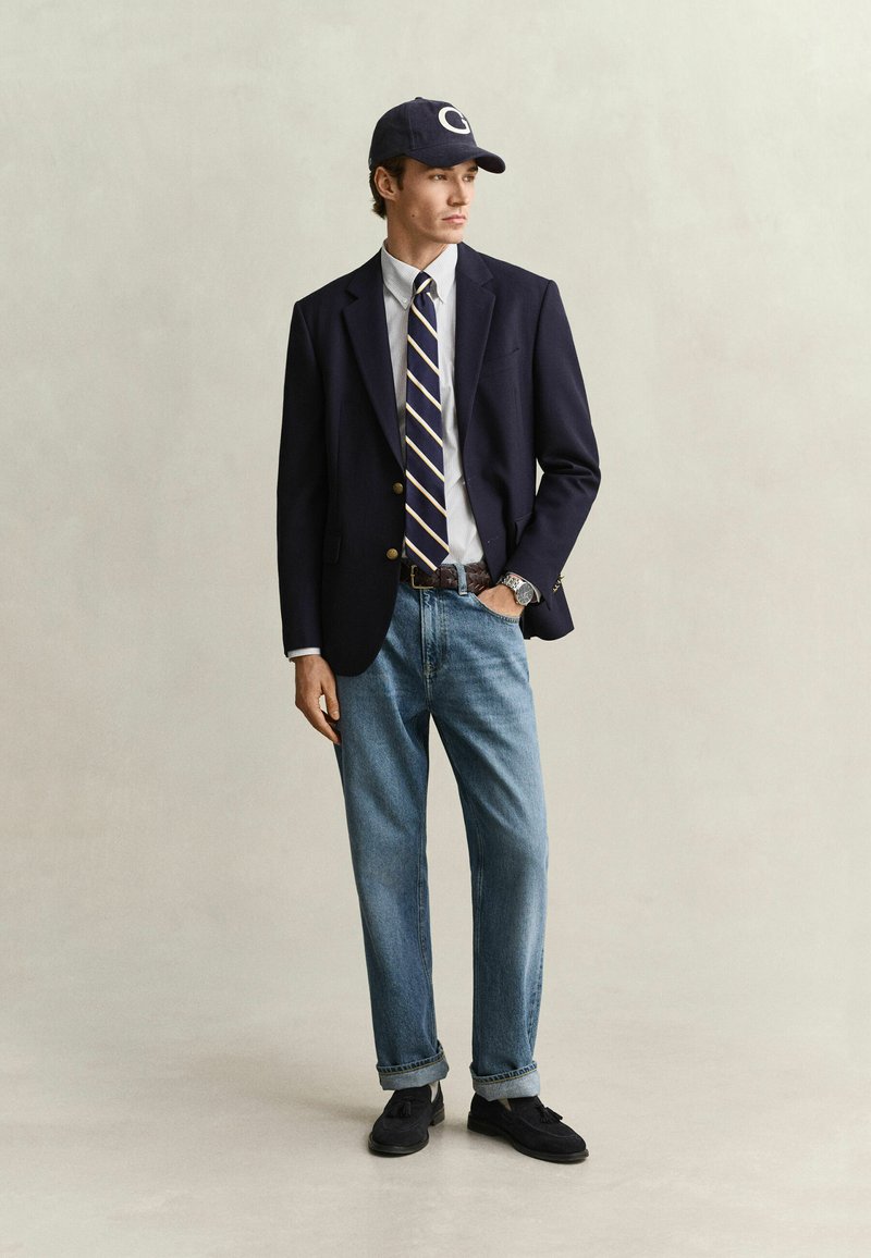 Young man wearing a navy blazer, white shirt, striped tie, blue jeans, navy cap, and black loafers standing with one hand in pocket.