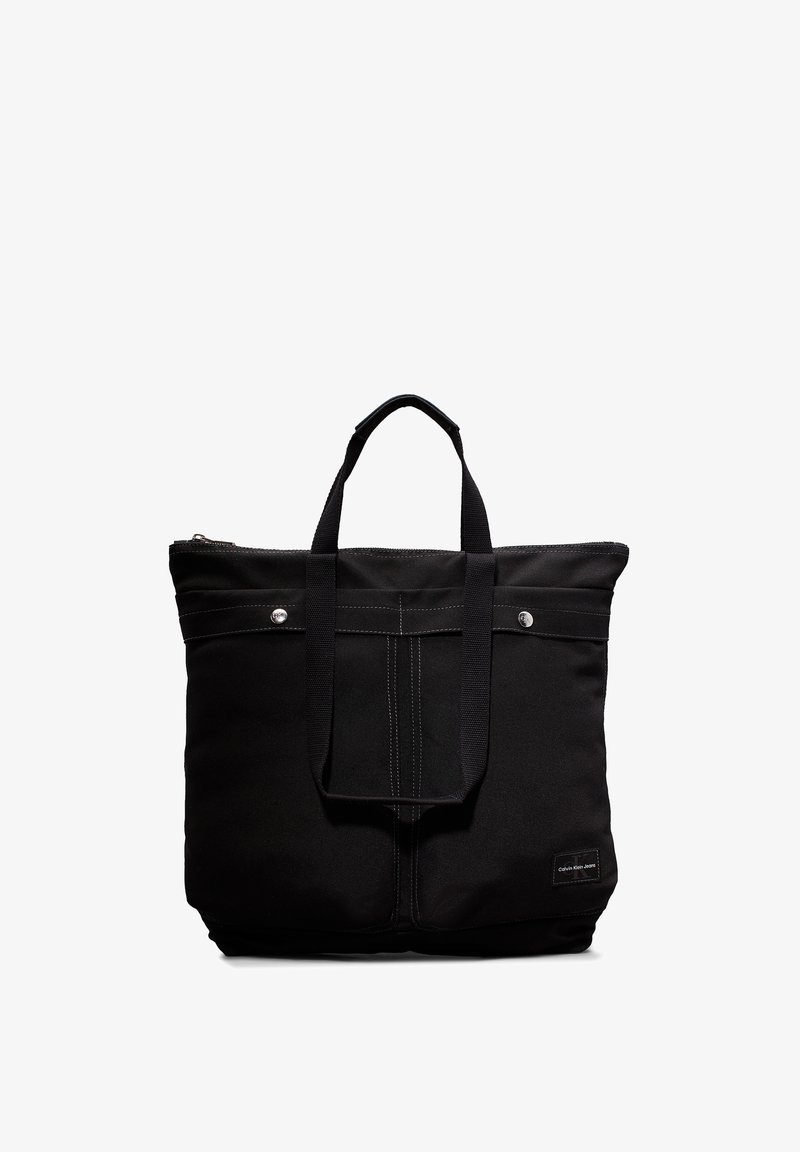 Svart stoff totebag med doble håndtak, glidelås og front sydd detalj. Har logo-patch og sølvfargede hardware-aksenter.