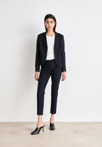 Blazer blu navale su misura con due bottoni, abbinato a pantaloni blu attillati e mule nere con tacco alto. Tessuto morbido con un design strutturato.