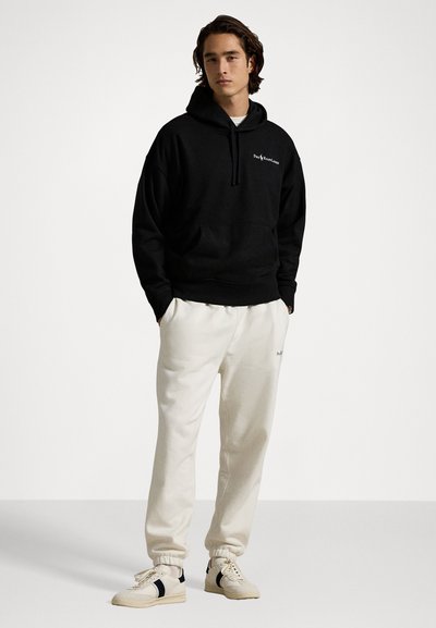 Zwarte hoodie met trekkoord en voorzak, gecombineerd met crèmekleurige joggers met boorden. Schoenen zijn beige met zwarte accenten.