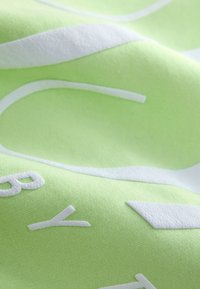 Zöld pamutszövet texturált felülettel, fehér vastag betűkkel és formákkal. Simaságot, egységes színt és modern grafikai kialakítást mutat.