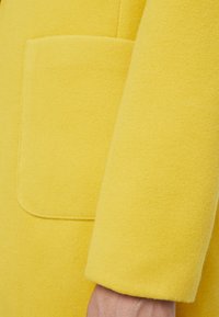 Manteau en laine jaune avec une texture douce, doté d'une grande poche à rabat et d'un poignet arrondi sur la manche.