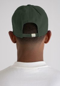 Rear view of a person wearing a dark green baseball cap and a white crew-neck shirt against a plain background.