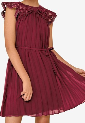 Donna che indossa un vestito plissettato bordeaux senza maniche con dettagli in pizzo sulle spalle e una sottile cintura legata in vita.