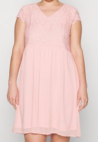 Kvinna klädd i en knälång rosa klänning med korta ärmar i spets och V-ringning, stående mot en enfärgad bakgrund.