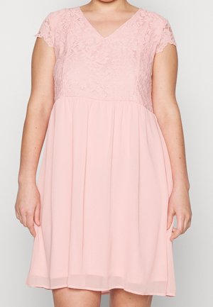 Vrouw in een roze jurk tot de knie met kant korte mouwen en een V-hals, staande tegen een effen achtergrond.