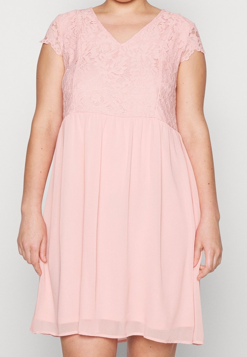 Kvinna klädd i en knälång rosa klänning med korta ärmar i spets och V-ringning, stående mot en enfärgad bakgrund.