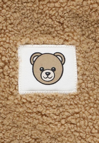 Tessuto marrone testurizzato con un'etichetta tessuta che presenta il volto di un orso cartone. L'orso ha una combinazione di colori marrone chiaro e crema con occhi neri.