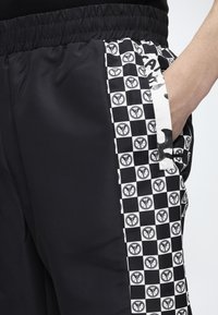 Μαύρο παντελόνι με ελαστική μέση, με ασπρόμαυρες καρό πλαϊνές λωρίδες με στρογγυλά λογότυπα και ένα χέρι ακουμπισμένο στην τσέπη.