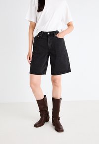 Black denim shorts with a high waist, cut above the knee. Paired with brown knee-high boots featuring buckles, set against a light background.