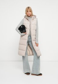 Vesta padded bej deschis cu glugă, cu deschidere în față și croială slim. Asortată cu un top gri cu mâneci lungi și pantaloni din denim deschis cu tăietură evazată. Ține o geantă neagră.