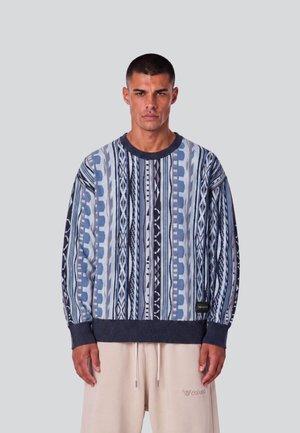 Mann trägt einen blau-grau gemusterten Langarmpullover und beigen Hosen und steht vor einem einfarbigen hellgrauen Hintergrund.