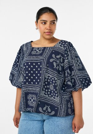 Kvinde med mørkt hår har en navyblå bluse med puffede ærmer og hvide paisley- og blomsterpatchworkmønstre, kombineret med lyseblå jeans.