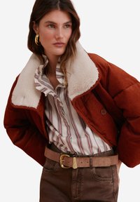 Hnědooranžová kordová bunda s krémovým beránkovým límcem, kombinovaná s pruhovanou košili a hnědým páskem. Má zapínání na knoflíky a jemnou strukturu.