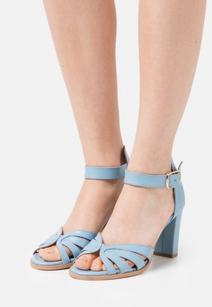 Light blue heeled sandals with an ankle strap, featuring a cut-out design on the toe area and a sturdy block heel. Smooth leather texture.
