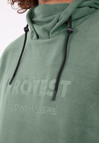 Groene fleece hoodie met een hoge kraag, voorzien van zwarte trekkoorden en gebosselde tekst "PROTEST SNOWCHASERS" op de borst. Zachte textuur.
