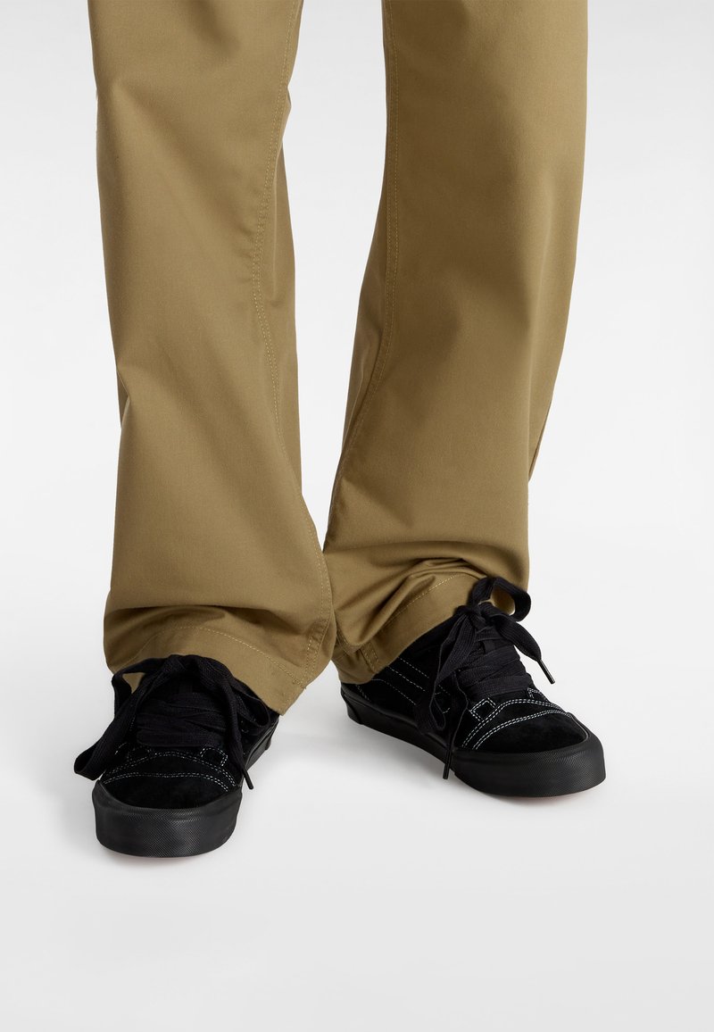 Sneaker nere con tomaie in suede e canvas, dotate di lacci tondi e una suola in gomma testurizzata, abbinate a pantaloni khaki.