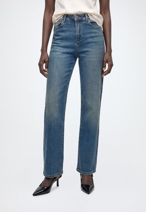 Person iført blå jeans med lige ben, en hvid top med blonder og sorte spidse højhælede sko, stående på en hvid baggrund.