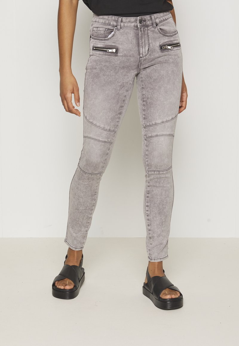 Jeans skinny cinzentos com bolsos com zíper, costuras nos joelhos e uma textura de lavagem desbotada. Usados com sandálias pretas com um design de tira larga.