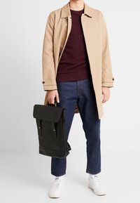 Uomo che indossa un cappotto beige, un maglione borgogna, jeans blu e sneakers bianche, mentre tiene uno zaino in pelle nera per il manico.