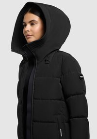 Schwarze Pufferjacke mit großer Kapuze, gesteppter Textur, Reißverschluss und Eingrifftaschen. Mit Logo auf dem linken Ärmel.