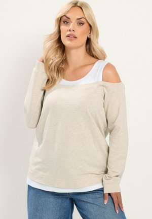 Blondynka w beżowym swetrze z odkrytymi ramionami nałożonym na biały top i niebieskie jeansy, pozująca na jednolitym jasnym tle.