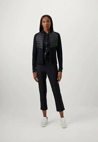 Donna in piedi che indossa una giacca nera con zip e pannelli imbottiti sul petto, pantaloni neri e sneaker bianche su uno sfondo semplice e chiaro.