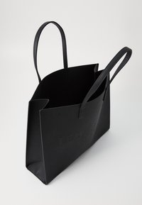 Sac fourre-tout noir avec des poignées allongées, fabriqué en matériau texturé. Présente le nom de la marque en relief à l'avant et une structure ouverte.