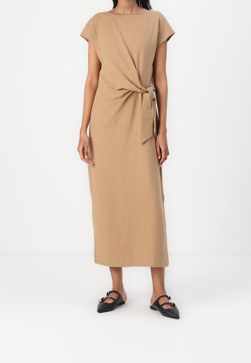 Femme portant une robe midi beige à manches courtes avec un détail de nœud sur le côté, associée à des chaussures plates pointues noires avec boucles.
